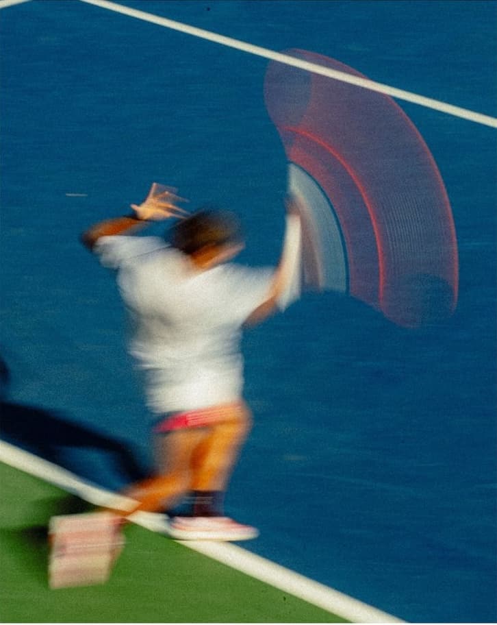 A grainy motion blur of a tennis player mid-swing on a blue court, with a red-orange racket trail.